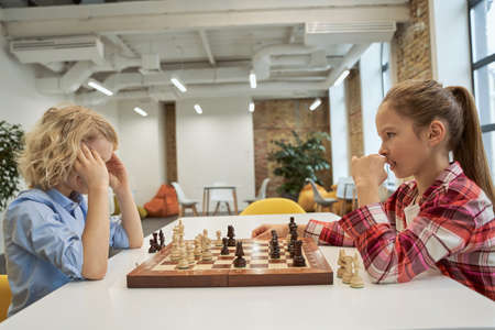Determination. Concentrated boy and girl looking at each other while playing chess, sitting together at the table in schoolの写真素材