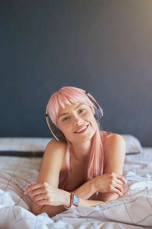 Portrait of beautiful model with pink hair listening to music on comfortable bed at home