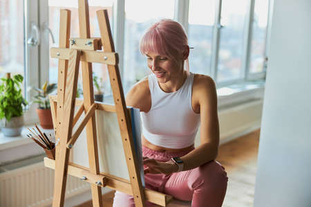 Smiling woman with pink hair draws on canvas at wooden easel on balconyの写真素材