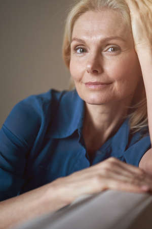 Portrait of peaceful mature woman looking at camera with friendly smile while relaxing on sofa at homeの写真素材