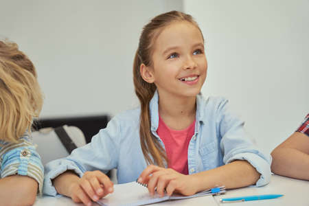 Portrait of adorable little schoolgirl smiling away and listening while studying, sitting at the table in elementary school classroomの写真素材