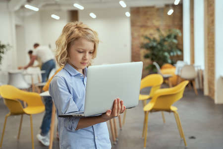Portrait of busy little boy holding and using laptop while posing in a classroomの写真素材