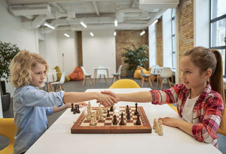 Respect. Adorable little friends, caucasian boy and girl shaking hands after match, playing chess, sitting together at the table in schoolの写真素材