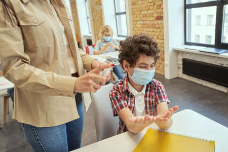 Little school boy wearing protective mask ready for cleaning his hands. Female teacher using an alcohol spray to disinfect student hands in classroomの写真素材