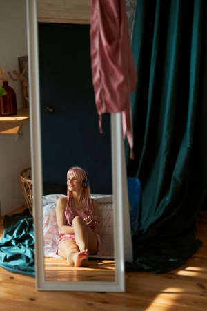 Dreamful pink haired lady listens to music reflecting in large mirror in bedroomの写真素材