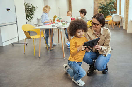 Full length shot of kind young female teacher smiling while showing video on tablet pc to her little student. Kids working on diy robot in the backgroundの写真素材