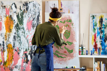 Back view of young female artist in apron painting picture, standing in living room at homeの写真素材