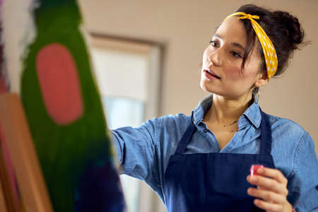 Portrait of young woman in apron looking inspired while working on abstract painting at cozy workshopの写真素材