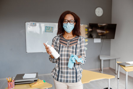 Woman in face mask cleaning the tables with antiseptic sanitizer in schoolの写真素材