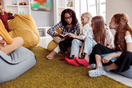 Teacher in glasses sitting on a pouf and reading for children in classの写真素材