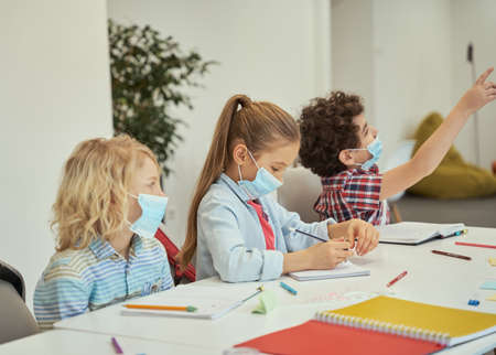 Little girl wearing protective mask looking at her notebook. Elementary school kids listening to teacher while sitting together at the table in a classroomの写真素材
