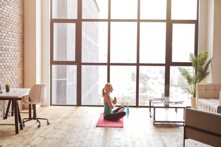 Side view of sportive mature woman sitting on a mat and watching online video tutorial using laptop while practicing yoga at home in modern apartmentの写真素材