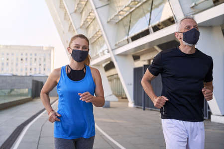 Sporty middle aged couple, man and woman wearing protective face masks running together in urban environment during coronavirus outbreakの写真素材