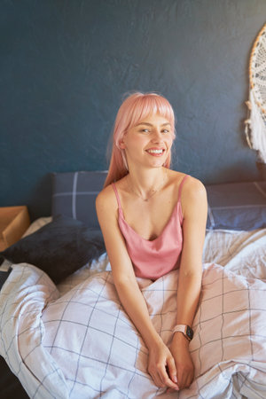 Cheerful woman with colorful hair in pink silk pajamas sits on bed in lazy morningの写真素材