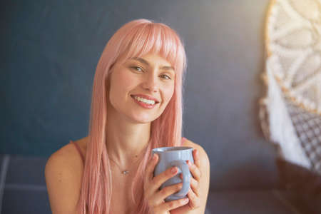 Portrait of stylish lady with dyed pink hair and mug against blue wall with decorの写真素材