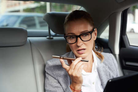 Multitasking. Portrait of a confident busy business woman using digital tablet and smartphone, recording voice message while sitting on back seat in the carの写真素材