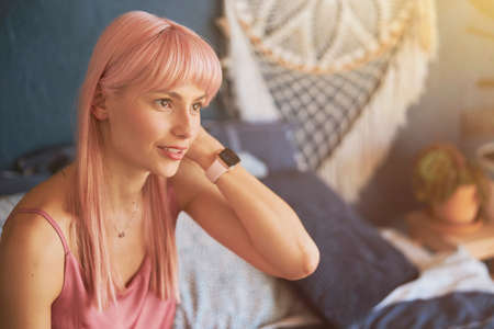 Elegant woman with pink hair and modern smartwatch sits on bed with pillowsの写真素材