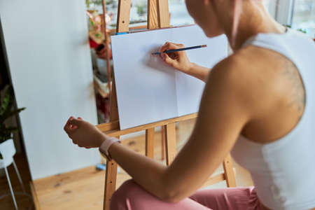 Woman artist draws picture on white paper with graphite pencil at wooden easel in light studioの写真素材