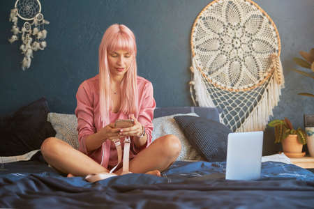 Woman with long pink hair uses mobile phone sitting near laptop on large bedの写真素材