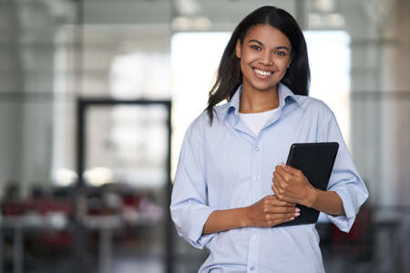 Portrait of a young cheerful beautiful business woman holding digital tablet, looking at camera and smiling while working in the modern officeの写真素材