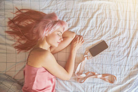 Beautiful sleeping female model with pink hair lies with mobile phone on large bedの写真素材