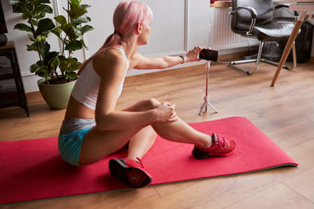 Athletic lady with pink hair turns on camera on smartphone to shoot video at homeの写真素材