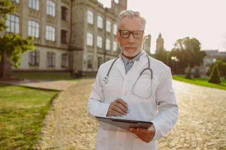 Portrait of concentrated senior male doctor in lab coat and glasses holding clipboard, looking through notes, standing outdoors near clinicの写真素材