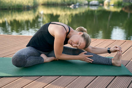 Flexibility. Relaxed middle aged woman sitting in yoga pose, stretching her body while doing yoga on a mat in a green parkの写真素材