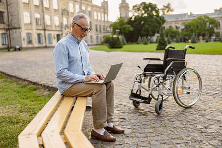 Full length shot of mature man, recovering patient with wheelchair working on a laptop while sitting on the bench in the park near clinicの写真素材