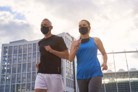 Motivated middle aged couple in protective face masks jogging together in urban environment in the morningの写真素材