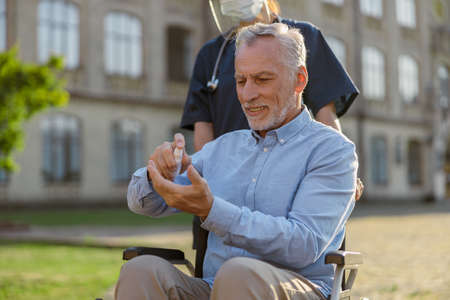 Senior man in wheelchair cleaning his hands with antibacterial sanitizer as a hygiene measure to prevent Covid19 infection while walking assisted by nurse outdoorsの写真素材