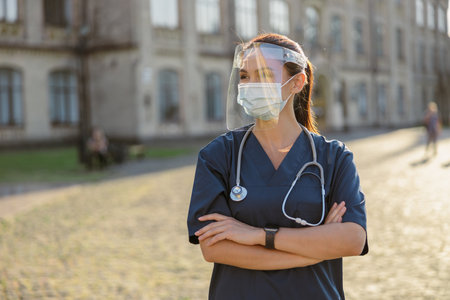 Young woman nurse wearing face shield and mask for safety which is required to prevent Covid19, standing outdoors on a sunny dayの写真素材