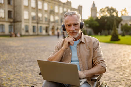 Portrait of happy mature man, recovering patient in wheelchair wearing headphones smiling at camera, using laptop outdoors in the park near clinicの写真素材