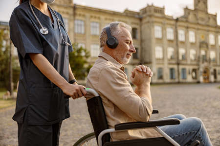Dreamy mature handicapped man in wheelchair wearing headphones and listening to music with eyes closed outdoors on a walk with nurseの写真素材