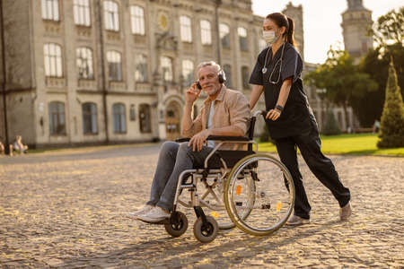Young female care taker in protective mask spending time with senior man in wheelchair wearing headphones and listening to music outdoors in the park near clinicの写真素材