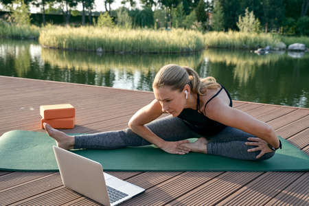 Discover yoga. Sporty attractive middle aged woman watching tutorial on a laptop while doing yoga on a mat in nature on a sunny dayの写真素材