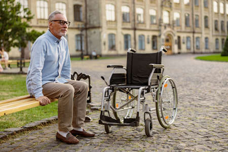 Full length shot of mature recovering patient with wheelchair sitting on the bench in the park near clinicの写真素材