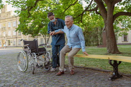 Full length shot of supportive young nurse wearing face shield and mask helping mature man, recovering patient in wheelchair to sit on the bench in the park near hospitalの写真素材