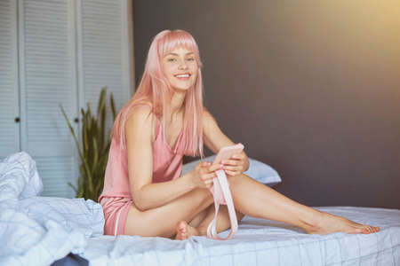 Beautiful lady with long pink hair holds smartphone sitting on comfortable bedの写真素材