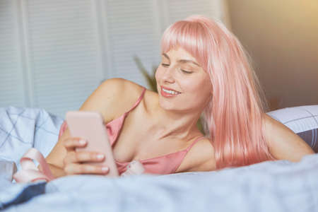 Beautiful woman texts on mobile phone leaning on hand in comfortable bed atの写真素材