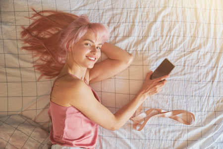 Cheerful lady with pink hair holds mobile phone with blank screen on comfortable bedの写真素材