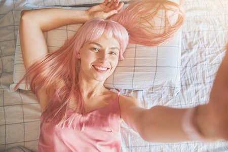 Positive woman with colorful pink hair takes selfie lying on bed with checkered linenの写真素材