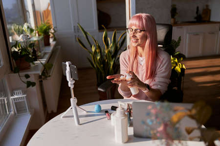 Cheerful woman with glasses applies cream onto palm from bottle shooting tutorial at tableの写真素材