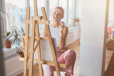Positive woman with colorful hair bites pencil sitting at easelの写真素材