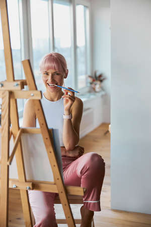 Attractive woman with pink hair bites pencil at easel on balconyの写真素材