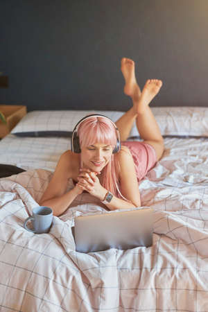 Pretty female model with headphones and cup looks at screen of modern laptop on bedの写真素材