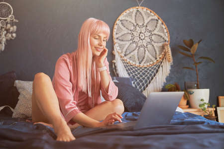 Attractive woman wearing silk gown surfs internet with laptop on large bed by dark blue wallの写真素材
