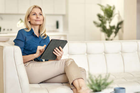 Relaxed mature lady in casual wear looking away, using tablet pc while sitting on a couch in modern apartment on a daytimeの写真素材