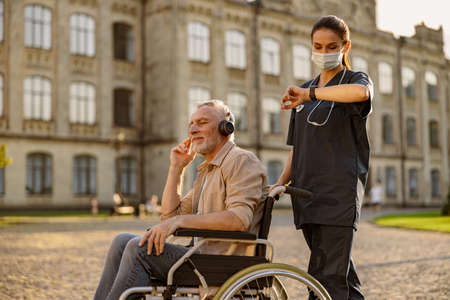 Young female care taker in protective mask spending time with senior man in wheelchair wearing headphones and enjoying music outdoors in the park near clinicの写真素材