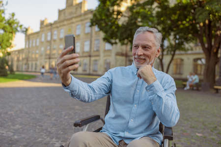 Smiling senior man, recovering male patient in wheelchair making a video call using smartphone while spending time alone in the park near hospitalの写真素材